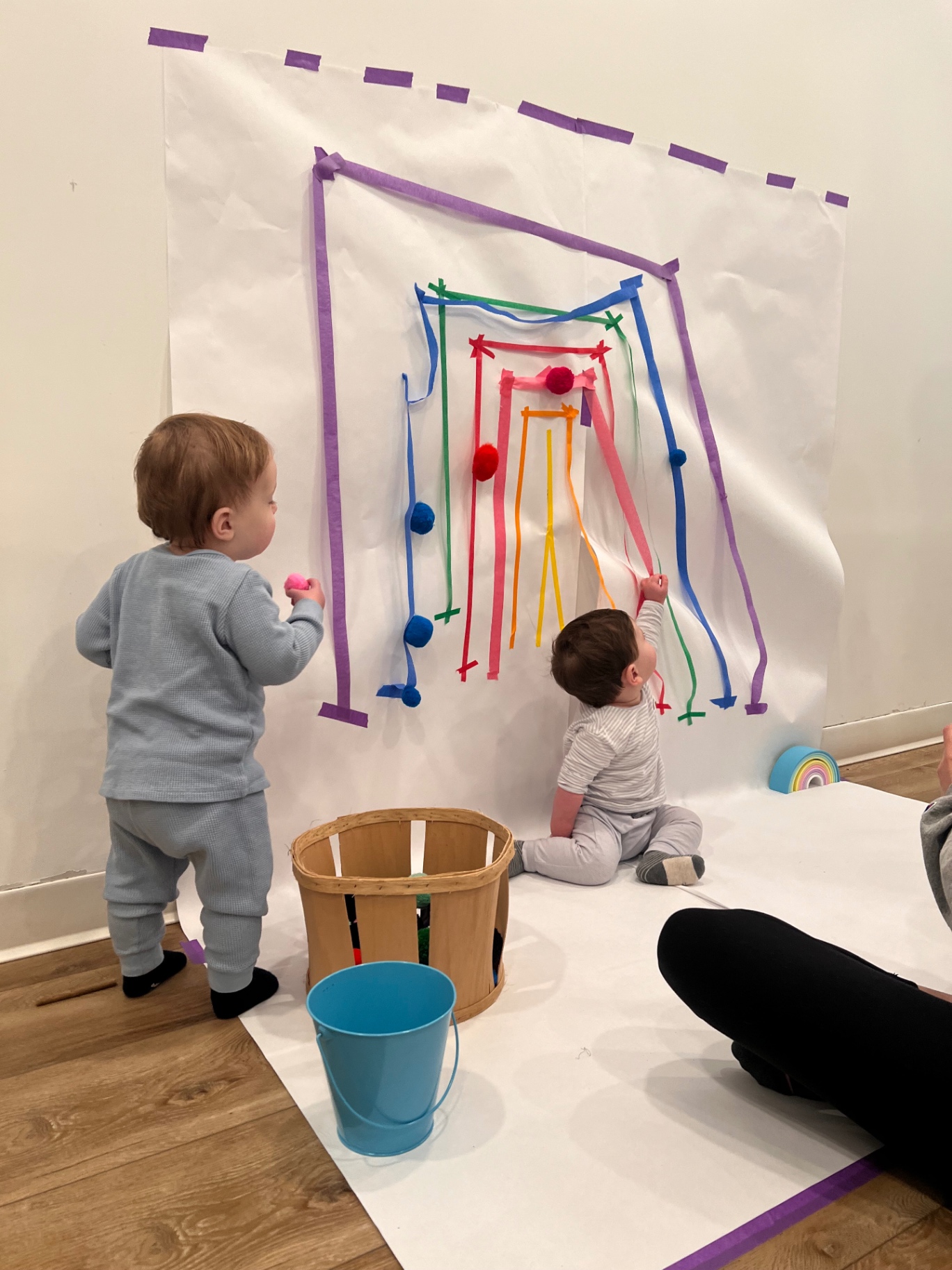 Toddlers exploring a rainbow tape and pom-pom wall